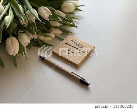 Sticky note with Women's day handwritten congratulations and white tulips bouquet laying at white office table. Greetings for women at workplace. 136366226