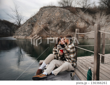 Couple enjoys time together by the water on Valentines Day while sharing warm drinks and smiles, wrapped in cozy sweaters 136366261