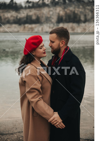 Couple shares a romantic moment by the water on Valentines Day in winter coats and a red beret 136366311