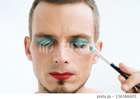 portrait of adult gay man with makeup and cosmetics on face. A handsome male with red lipstick applied eye shadow sparkle on his eyes with brush 136366605