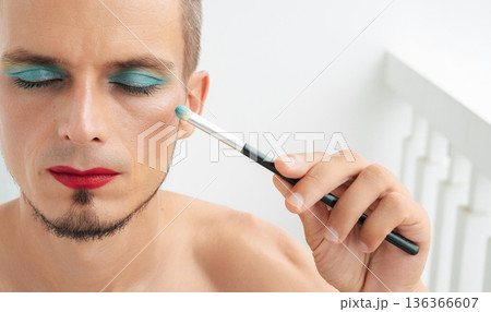 portrait of feminine gay man with makeup and cosmetics on face. A handsome young transgender male with red lipstick applied eye shadow sparkle on his eyes with a brush on a white background portrait of feminine gay man with makeup and cosmetics on face. A handsome young transgender male with red lipstick applied eye shadow sparkle on his eyes with a brush on a white background 136366607