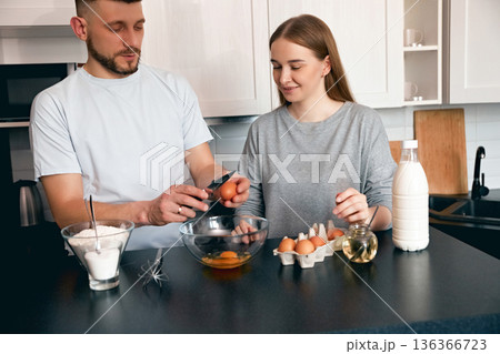 Couple prepares breakfast in a kitchen with eggs, milk, and flour on a counter while smiling and working together 136366723