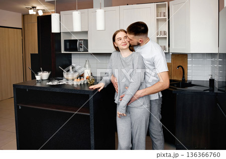 Couple enjoys cooking together in a modern kitchen during the evening while preparing a meal and sharing moments of affection near the counter Couple enjoys cooking together in a modern kitchen during the evening while preparing a meal and sharing moments of affection near the counter 136366760