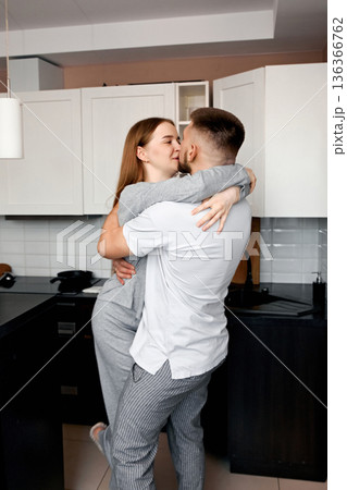 Couple shares a kiss in a kitchen, enjoying time together while embracing each other in a cozy home setting during the evening 136366762