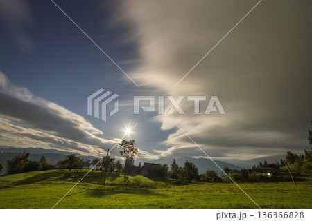 Summer night mountain panorama. Old wooden weathered shepherd huts on green clearing on cloudy evening sky background, bright road and dwelling lights deep below in valley. Summer night mountain panorama. Old wooden weathered shepherd huts on green clearing on cloudy evening sky background, bright road and dwelling lights deep below in valley. 136366828