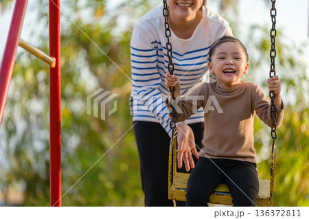cheerful child girl with mother playing on swing at playground 136372811