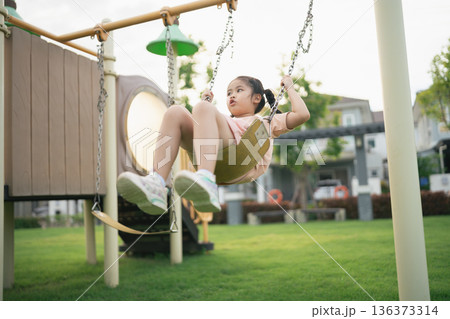 Young girl enjoying a sunny day on a swing in a playground, surrounded by vibrant greenery and modern architecture, capturing childhood joy and innocence 136373314