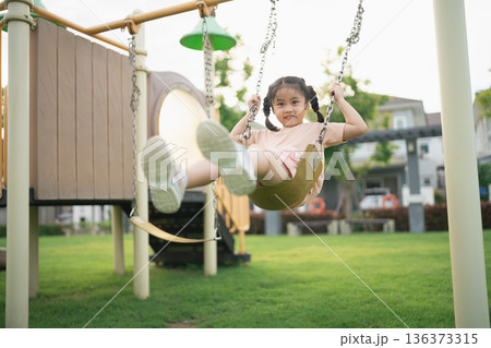 Young girl enjoying playful moments on swing set in vibrant park setting with bright sunshine and green landscape, capturing joy and childhood innocence 136373315