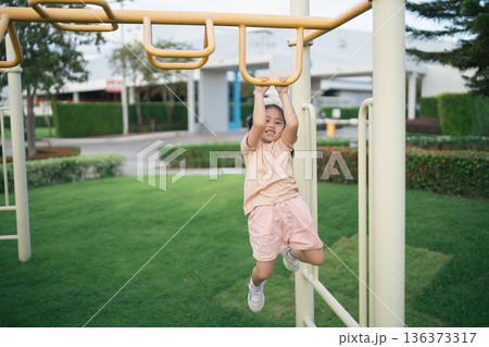 Happy child swinging on monkey bars in a playground, enjoying outdoor playtime and laughter during a sunny afternoon in a park setting Happy child swinging on monkey bars in a playground, enjoying outdoor playtime and laughter during a sunny afternoon in a park setting 136373317