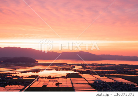 滋賀県東近江市猪子町の猪子山から望む水張りの田植え前の水田がオレンジ色に光る夕景 滋賀県東近江市猪子町の猪子山から望む水張りの田植え前の水田がオレンジ色に光る夕景 136373373