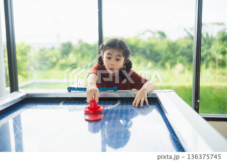 Young girl playing air hockey indoors with concentration and excitement, natural light streaming through large windows, green landscape in background Young girl playing air hockey indoors with concentration and excitement, natural light streaming through large windows, green landscape in background 136375745