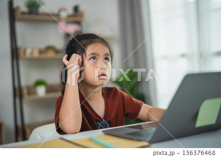 Young girl with frustrated expression sitting at a desk with laptop and stationery during a study session at home, showcasing student struggles and concentration issues 136376468