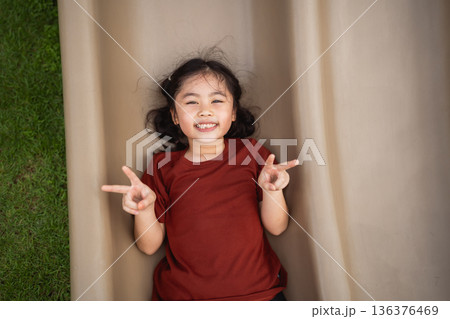 Joyful child sliding down playground slide, smiling with peace signs, enjoying outdoor playtime, surrounded by green grass in bright sunlight 136376469