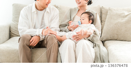 家のソファに座りで赤ちゃんに哺乳瓶で水を飲めせる母親,ママ,家族 136376478