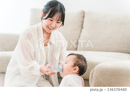 家のリビングで赤ちゃんに哺乳瓶で水を飲めせる母親,ママ 136376480