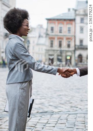 Two professional women in business attire shake hands on a cobblestone street with historic buildings in the background, symbolizing agreement and partnership Two professional women in business attire shake hands on a cobblestone street with historic buildings in the background, symbolizing agreement and partnership 136379224