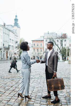 Two professionals, a man and a woman, shake hands on a cobblestone street in front of historic buildings Two professionals, a man and a woman, shake hands on a cobblestone street in front of historic buildings 136379228