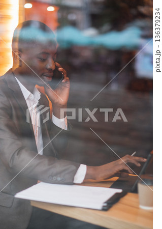 A businessman in a suit talks on his mobile phone while working on a tablet at a wooden table with papers and a coffee cup A businessman in a suit talks on his mobile phone while working on a tablet at a wooden table with papers and a coffee cup 136379234