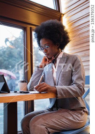A focused Black woman in a suit writes in a notebook at a table in a cafe, with a tablet and coffee cup nearby 136379352