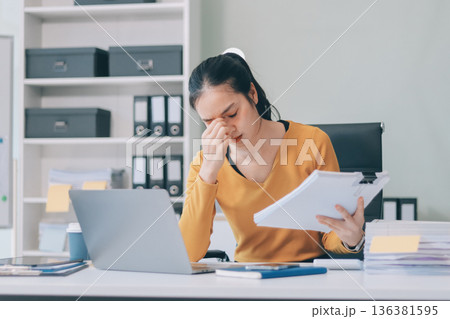 Young professional woman suffering from headache while analyzing financial charts and graphs on computer, experiencing work related stress in modern office Young professional woman suffering from headache while analyzing financial charts and graphs on computer, experiencing work related stress in modern office 136381595
