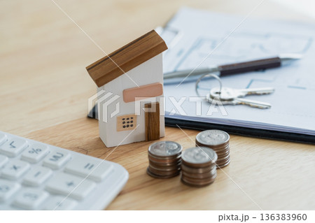 Miniature wooden house model on desk with coins, calculator, keys, and financial documents illustrating real estate investment concept and home buying decision 136383960