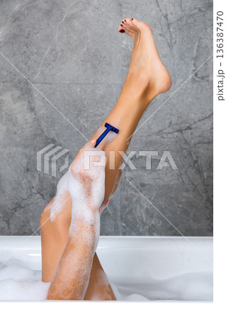 Woman shaving legs with razor in bathtub on gray wall background. Woman shaving legs with razor in bathtub on gray wall background. 136387470