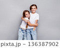 Happy children brother and sister hugging and smiling in studio, joyful siblings in casual white t-shirts on clean grey background, beautiful kids model together 136387902