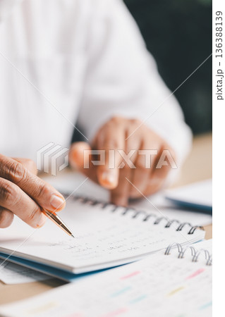 Hand writing notes in a notebook beside a calculator on office desk for budgeting and accounting. Personal finance planning, expense tracking, tax calculation, soft focus background, copy space. 136388139