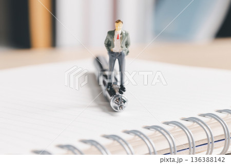 Miniature businessman standing on a pen over a spiral notebook, symbolizing leadership and planning. Business strategy, meeting notes, office productivity, consulting concept with copy space. Miniature businessman standing on a pen over a spiral notebook, symbolizing leadership and planning. Business strategy, meeting notes, office productivity, consulting concept with copy space. 136388142