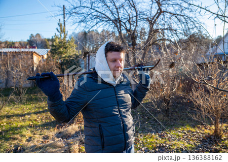 young Caucasian male farm worker with rake on a sunny autumn day in orchard. Farm work, harvest, cleaning, or gardening concept 136388162