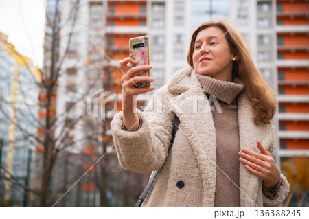 Caucasian Woman takes selfie photo with smartphone, surfs news, writes message on social media. She is surrounded by modern buildings and trees in the background. Caucasian Woman takes selfie photo with smartphone, surfs news, writes message on social media. She is surrounded by modern buildings and trees in the background. 136388245