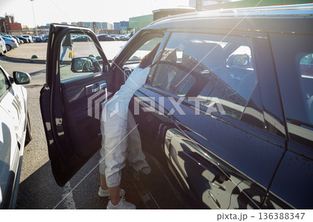 Woman bending into a car on a parking lot getting items from seat. Daily errands and vehicle convenience concept. Shopping trip to the mall Woman bending into a car on a parking lot getting items from seat. Daily errands and vehicle convenience concept. Shopping trip to the mall 136388347