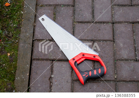 Hand saw with red and black handle on a brick path, detail of a sharp blade and teeth for cutting, gardening tool. 136388355