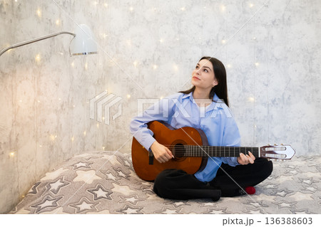 actress, singer, influencer, blogger. Woman playing acoustic guitar while sitting on bed, looking up thoughtfully with sparkling wall decorations, musical inspiration and dream 136388603