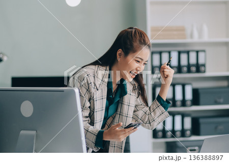 Determined asian businesswoman works late in a modern office, confidently analyzing data on her laptop, embodying success and professionalism with a smile 136388897