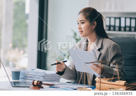 Confident Lawyer Working at Desk: A young Asian female lawyer confidently works on her laptop, surrounded by legal symbols. She exudes professionalism and competence in her modern office setting. 136388935