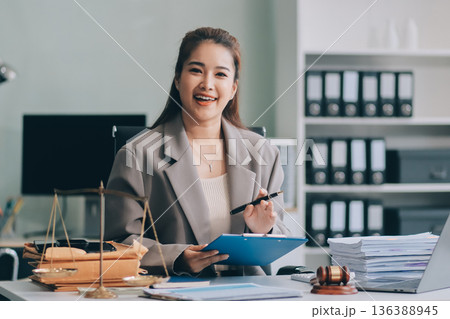 Confident Lawyer Working at Desk: A young Asian female lawyer confidently works on her laptop, surrounded by legal symbols. She exudes professionalism and competence in her modern office setting. 136388945