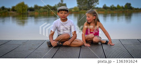A group of cute little children sit on a wooden pier by a lake. Small children looking into the future. Children sit on a dock. Siblings. Two children of different ages a school-age boy and a A group of cute little children sit on a wooden pier by a lake. Small children looking into the future. Children sit on a dock. Siblings. Two children of different ages a school-age boy and a 136391624