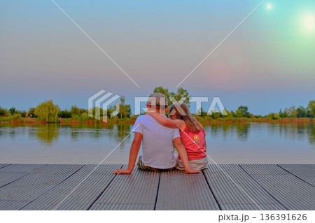 A little boy wearing a flat cap and a cute little girl sit in each other's arms on a wooden pier by the lake. Small children sit on a pier at the edge of a pond. The concept of a carefree childhood A little boy wearing a flat cap and a cute little girl sit in each other's arms on a wooden pier by the lake. Small children sit on a pier at the edge of a pond. The concept of a carefree childhood 136391626