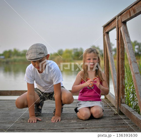 Children sitting on pier. Two children of different age - elementary age boy and preschool girl sitting on a wooden pier. Girl making heart shape. Summer and childhood concept. Children on bench at 136391630