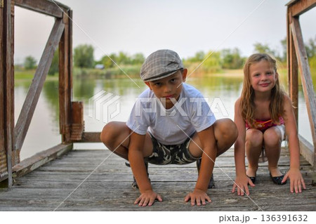 A little boy and sweet girl are sitting on a wooden pier on the bank of a lake. Small children are sitting on a pier at the side of a pond. The concept of a carefree childhood and summer holidays. 136391632