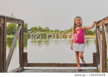 A small smiling girl stands on a wooden pier at the side of a lake. The photo was taken during the golden hour, at sunset. It evokes the concept of childhood, summer vacation and carefree days. 136391639