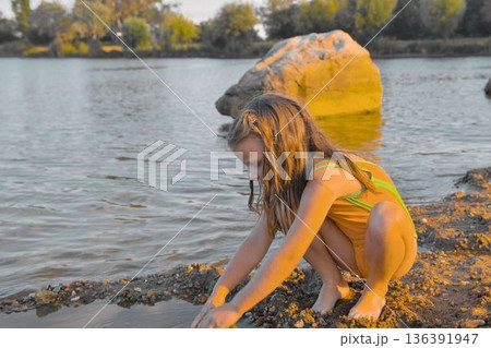 A little girl in a swimsuit plays in the sand at the edge of a pond. The concept of summer holidays, carefree days, and childhood. A little girl in a swimsuit plays in the sand at the edge of a pond. The concept of summer holidays, carefree days, and childhood. 136391947