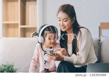 A cheerful Asian mother helps her young daughter do homework and color a book at home, while the father works on a laptop in the background. A cozy, bright, and modern family lifestyle. A cheerful Asian mother helps her young daughter do homework and color a book at home, while the father works on a laptop in the background. A cozy, bright, and modern family lifestyle. 136393900