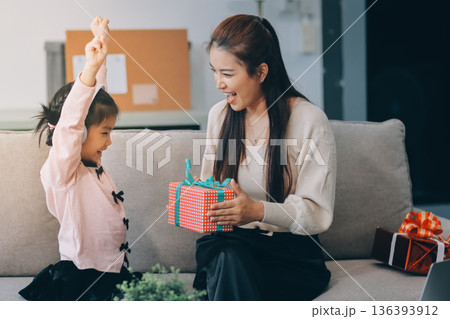 A cheerful Asian mother helps her young daughter do homework and color a book at home, while the father works on a laptop in the background. A cozy, bright, and modern family lifestyle. A cheerful Asian mother helps her young daughter do homework and color a book at home, while the father works on a laptop in the background. A cozy, bright, and modern family lifestyle. 136393912