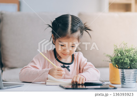 A cheerful Asian mother helps her young daughter do homework and color a book at home, while the father works on a laptop in the background. A cozy, bright, and modern family lifestyle. 136393923