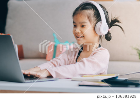 A cheerful Asian mother helps her young daughter do homework and color a book at home, while the father works on a laptop in the background. A cozy, bright, and modern family lifestyle. A cheerful Asian mother helps her young daughter do homework and color a book at home, while the father works on a laptop in the background. A cozy, bright, and modern family lifestyle. 136393929