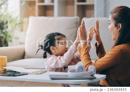 A cheerful Asian mother helps her young daughter do homework and color a book at home, while the father works on a laptop in the background. A cozy, bright, and modern family lifestyle. A cheerful Asian mother helps her young daughter do homework and color a book at home, while the father works on a laptop in the background. A cozy, bright, and modern family lifestyle. 136393935