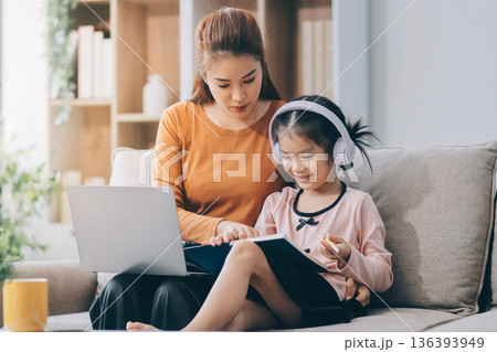 A cheerful Asian mother helps her young daughter do homework and color a book at home, while the father works on a laptop in the background. A cozy, bright, and modern family lifestyle. 136393949