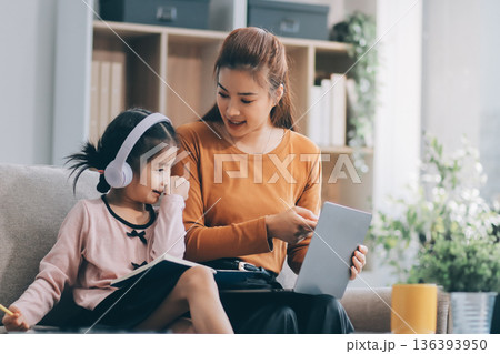 A cheerful Asian mother helps her young daughter do homework and color a book at home, while the father works on a laptop in the background. A cozy, bright, and modern family lifestyle. 136393950